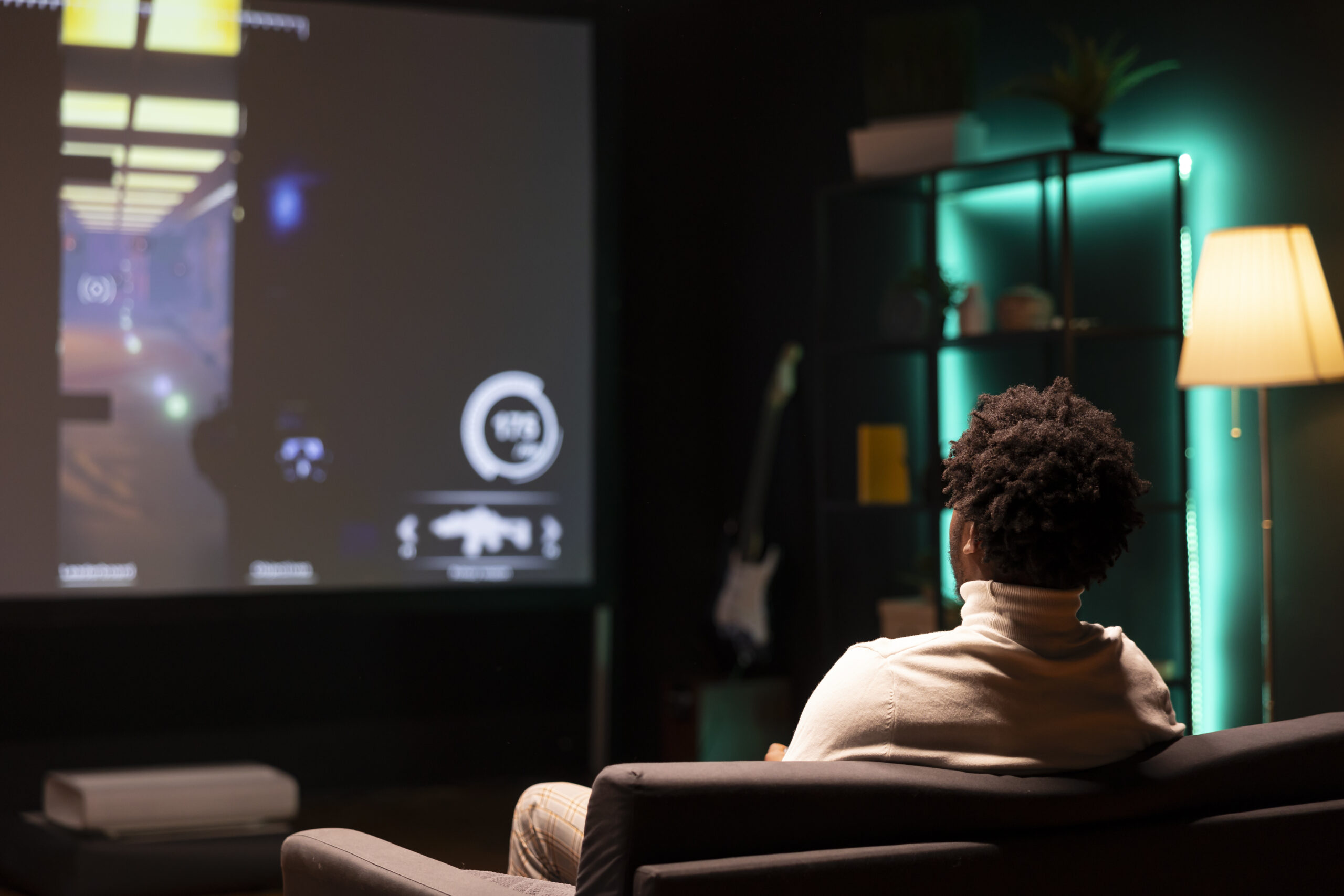Gamer in front of ultrawide smart TV, watching first person shooter videogame gameplay. African american man in home theatre using high tech television set to see gaming content online